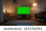 Small photo of Shot of a TV with Horizontal Green Screen Mock Up. Cozy Evening Living Room with a Chair and Lamps Turned On at Home.