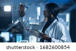 Small photo of In the Research Laboratory Smart and Beautiful African American Female Scientist Wearing White Coat and Protective Glasses Writes Formula on Glass Whiteboard, References Her Tablet Computer