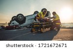 Small photo of On the Car Crash Traffic Accident Scene: Paramedics Saving Life of a Female Victim who is Lying on Stretchers. They Listen To a Heartbeat, Apply Oxygen Mask and Give First Aid. Background Firefighters
