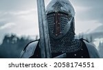 Small photo of Medieval Knight Wearing Armour and Helmet, Draws Sword from Shearh, Ready to Fight, Kill His Enemy in Battle. Warrior Soldier on Battlefield. War, Invasion, Crusade. Cinematic Historic Reenactment