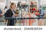 Small photo of Beautiful Smiling Female Customer Shopping in Clothing Store, Using Smartphone, Browsing Online, Comparing on Internet, Choosing Stylish Clothes. Fashionable Shop, Colorful Brands, Sustainable Designs