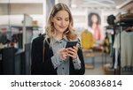 Small photo of Beautiful Smiling Female Customer Shopping in Clothing Store, Using Smartphone, Browsing Online, Comparing on Internet, Choosing Stylish Clothes. Fashionable Shop, Colorful Brands, Sustainable Designs