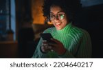 Small photo of Cinematic Portrait of Black Female with Curly Hair Sitting on the Floor at Home and Using Smartphone. Diverse Woman Receives Good News and Chatting with Friends Via Social Media.
