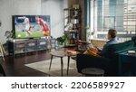Small photo of Sports Fan Watches Important Soccer Match on TV at Home, He Aggressively Gestures with the Fist, Cheering for His Team. His Successful Team Scores a Goal and He Celebrates the Win.