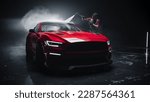 Small photo of Adult Car Washer in Uniform Washing a Red Performance Car with a High Pressure Cleaner. Cleaning Technician Working on a Stylish American Car in a Dark Room. Commercial Studio Footage for Advertising