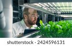 Small photo of Portrait of Agricultural Grower Working in a Corridor in Vertical Farm Facility Next to a Rack with Freshly Grown Plants. Caucasian Hydroponics Technician Closely Studying and Cultivating Crops.