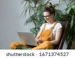 Young female gardener in glasses wearing overalls, sitting on wooden chair in greenhouse, using laptop after work, communicates on Internet with customer. Plant on background.Home gardening, freelance