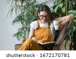 Small photo of Young female gardener in glasses wearing overalls, resting after work, sitting on wooden chair in home greenhouse, holding reusable foldable coffee/tea mug, smiling and reading book on her knees.