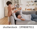 Small photo of Naughty kid rumbling kitchenware while tired mother try to sleep on sofa in living room. Hyperactive preschool son make noises disturb young mom stressed and annoyed. Motherhood and children concept