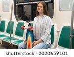 Millennial woman with long hair holding longboard sitting in subway train, smiling, looking at camera. Portrait of happy European young girl skateboarder with skate board in metro train. Urban hobby.