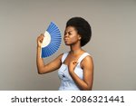 Small photo of Young afro woman suffering from heat or high air temperature inside, waving with paper fan, standing isolated on grey. Overheated african american female fanning herself to cool down on hot summer day