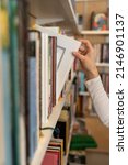 Small photo of Female hand pulling book from bookshelf in public library in university, college or high school. Woman student take novel from bookcase in bookshop store, soft focus. Education and literature concept