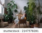 Small photo of Overworked woman freelancer sitting on chair in cozy greenhouse, resting, stretching arms with closed eyes. Smiling tired woman in home garden taking break from online study. Plant lovers concept.