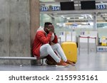 Small photo of Sad African American man upset at airport his flight is delayed. Depressed traveler male waiting for a plane sitting in empty terminal with baggage. Exhausted guy on a long night connection at airport
