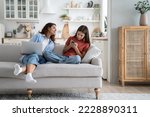 Small photo of Independent everyday teen girl keeps secrets from mom by hiding phone screen and avoiding surveillance. Smiling young woman with laptop on lap sits on sofa trying to make friends with daughter