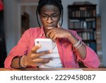 Small photo of Frightened anxious African American man reading news in mobile phone or university dropout message. Displeased Black guy in glasses and casual clothes sits at table indoors with smartphone in hand