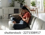 Unmotivated confused Asian woman sits with phone on sofa in apartment not willing to start working. Procrastinating Chinese student girl is addicted to mobile gadgets and Internet surfing 