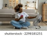 Summer heat at home. Happy curly African American woman sitting with smartphone on floor enjoying cool fresh air blowing from electric fan, chatting on mobile phone relaxing in front of air cooler