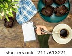 homemade chocolate muffins or madeleine in green plate with cup of coffee and pot of lilies flowers and empty notebook and woman holding pen on wooden table