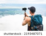 Back view of tourist man with backpack using smart phone on selfie stick shooting video blog. Asian traveler taking photo on vacation trip with beautiful sky on background.