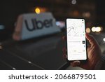 Small photo of Uber app displayed on smartphone held in hand in front of Uber taxi sign on top of a car at night with city lights background with soft focus. Taking a cab concept. Warsaw, Poland - October 23, 2021