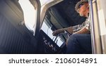 Small photo of Elderly Latino semi truck driver sits in the cab looking ahead wearing plaid shirt, sunglasses and hard hat. Handsome cool looking trucker sits at the steering wheel.