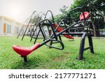 Small photo of Red seesaw in the playground. Playground equipment for children to play. Plastic seesaw or teeter-totter, swing, and slide at outdoor playground with green grass ground. Outdoor kids toy at park.