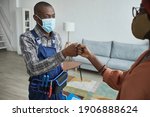 Small photo of Waist up portrait of African-American handyman wearing mask bumping fists with client as con tactless greeting, copy space