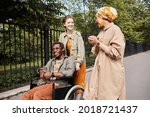 Young multi-ethnic friends talking during stroll: Caucasian woman pushing wheelchair with disabled man while talking to Arabian friend