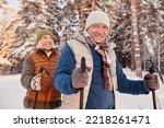 Waist up portrait of happy senior couple walking with poles in winter forest and smiling at camera