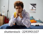 Cute serene teenage boy in casualwear keeping hands by chin while sitting on double bed in his bedroom at leisure and looking at camera