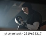 Small photo of Young Muslim female boxer in hijab putting on boxing gloves before fight with rival or training while standing in front of camera in dark gym