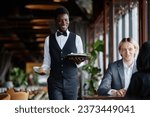 Small photo of Portrait of young Black man as waiter bringing food on tray to guests enjoying dinner in luxury restaurant
