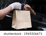 Small photo of Closeup food delivery man hand wear glove passing brown paper bag to woman customer driving thru pickup out of black car window meeting social distancing requirements and supporting small businesses