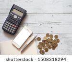 Smartphone white screen silver pen on blank page notebook with calculator and gold coins group on white wood table top view above space,Business concept
