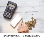 Smartphone white screen and hand holding silver pen on blank page notebook with calculator and gold coins group on white wood table top view above space,Business concept