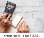 Hand with silver pen and smartphone press button of calculator on blank page diary on wooden table top view nature shadow copy space business concept