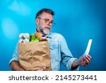 Small photo of Shocked mature man looking at store receipt after shopping, holding a paper bag with healthy food. Real people expression. Inflation concept. man with a paper bag of groceries looks surprised.