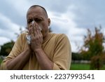 Small photo of Allergic middle aged man blowing on wipe in a park on spring season a sunny day. Sick Man With High Temperature is Using Handkerchiefs, Trying to Override the Sickness While Walking in Nature