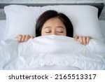Small photo of Cozy bedclothes. Asian girl hiding under blanket in bed, top view