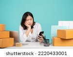 Small photo of Young disappointed puzzled sad Asian woman in casual shirt sit work at white office desk with pc laptop hold mobile cell phone prop up face isolated on pastel green background studio