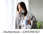 Small photo of Sick asian girl in medical face mask standing by the window and and cough, she being on quarantine with covid-19 or flu