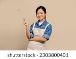 Small photo of Cheerful Asian woman wearing apron and holding hand whisk standing isolated on light brown background.