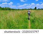 Woman riding bicycle single track trail running along a flood embankment among tall green grass in summer sunny day. Back view.