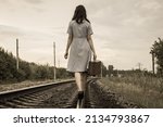 Small photo of A rear view of a young woman in a dress and with a suitcase walking away along the rails of a railway road. The concept of departure, emigration, departure from the country, refugee status,deportation