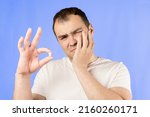 Small photo of Man in a white T-shirt on a blue background holds a wisdom tooth in his hands after surgical tooth extraction.Man after an operation to remove wisdom teeth.Pain in wisdom teeth, concept of dentistry