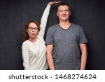 Studio waist-up shot of amazed short woman pulling up and showing with hand at height of tall man standing beside her, smiling and looking at camera, over gray background. Variety of person's heights