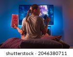 A blonde girl in pajamas watches TV at night. Insomnia. She is holding a pack of popcorn and a carbonated drink with a straw. Neon light. Watching TV shows and series, quarantine.