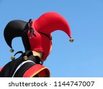 Small photo of Person in a jester costume isolated on clear blue sky, carnival concept. Actor in black-red foolscap with bells. Joker head close-up, back view