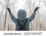 Woman wearing down jacket with hood standing with her hands raised and enjoying the snowy weather. Leisure in winter park, cold season